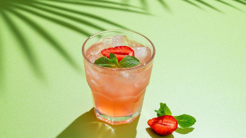 drinking glass of strawberry cocktail with mint leaves and iced cubes on bright green background, tropical tree shadow homemade ready to eat lemonade vitamin delicious refreshing drink, horizontal, copy space