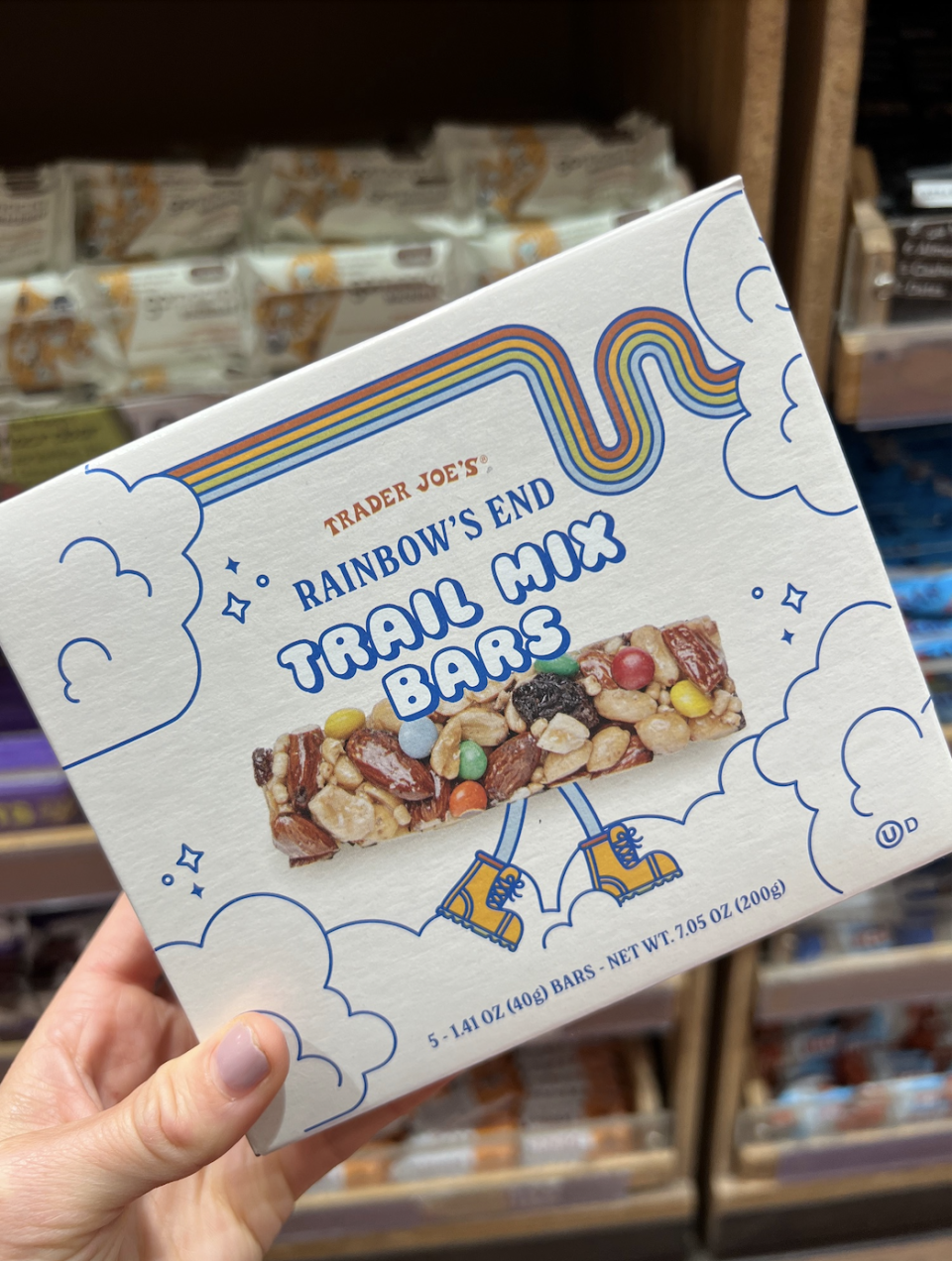 Hand holding a box of Trader Joe's Rainbow's End Trail Mix Bars in a store. The box features illustrations of clouds and a pot of gold