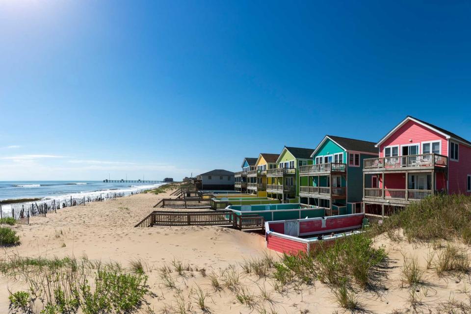 Side view of row of colorful beach houses in Nags Head. Pink house beginning the row, navy house last in row. Sand and Beach to left. Pier in background. People in pool at pink house.