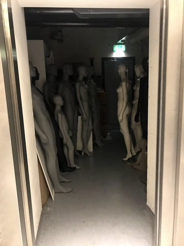 A dimly lit hallway filled with various standing mannequins of different sizes, all arranged closely together. Light source from the end of the hallway