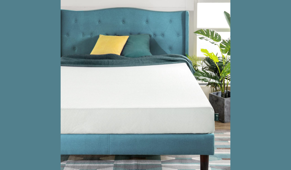 A white mattress lays on a large bed with a teal fabric headboard and multi-colored pillows 