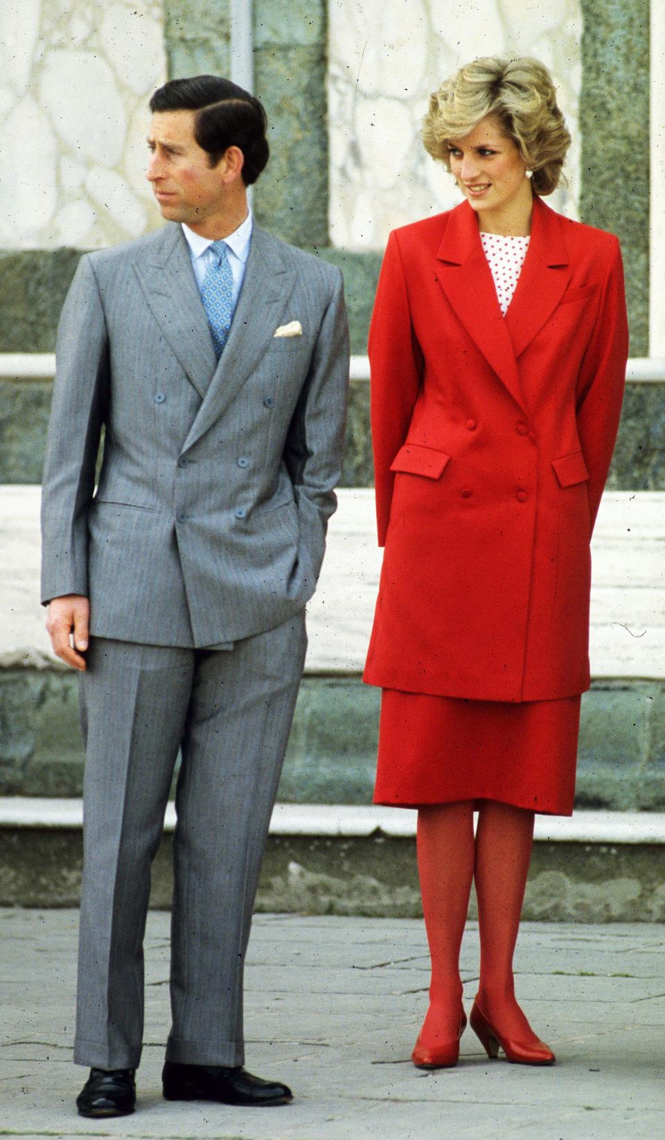 Princess Diana with Prince Charles in Florence. He is wearing a. grey mens suit, she is wearing a long red jacket, red skirt, red tights and red shoes, with a red and white polka dot top.