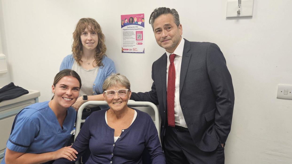 A group of four people in a white hospital room, a woman in blue scrubs leans towards a woman in a blue top who is sitting in a hospital chair, behind them are a man in a grey suit with a red tie and a woman in a blue cardigan with a taupe top underneath