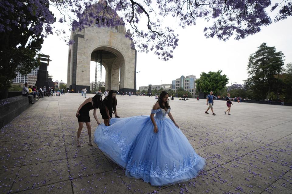 Two people adjust the train on a girl's blue gown, with a massive monument in the background and purple blooms overhead.