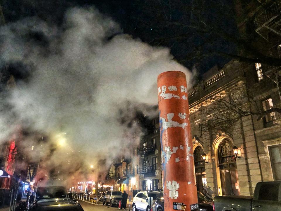 An orange upright pipe vents steam into the night sky in front of a building with stone arches and gaslamp-type lights, and parked cars on an urban street