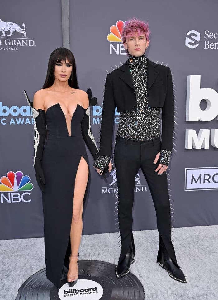 Megan Fox in a low cut black and silver gown, standing beside Machine Gun Kelly in a black and silver suit with spikes on the BBMAs red carpet.