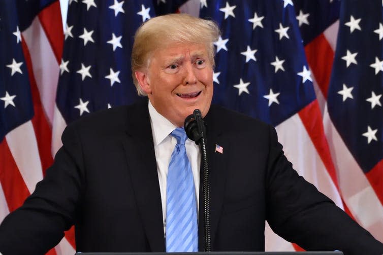 Donal Trump in a black suit and blue tie stands behind a lecturn in front of US flags and gestures in disbelief.