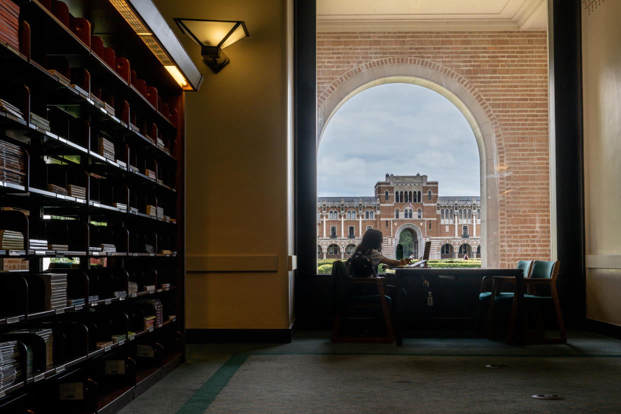 A woman sits at a table by a wall of bookshelves framed by a large brick archway, through which a large, stately building can be seen.