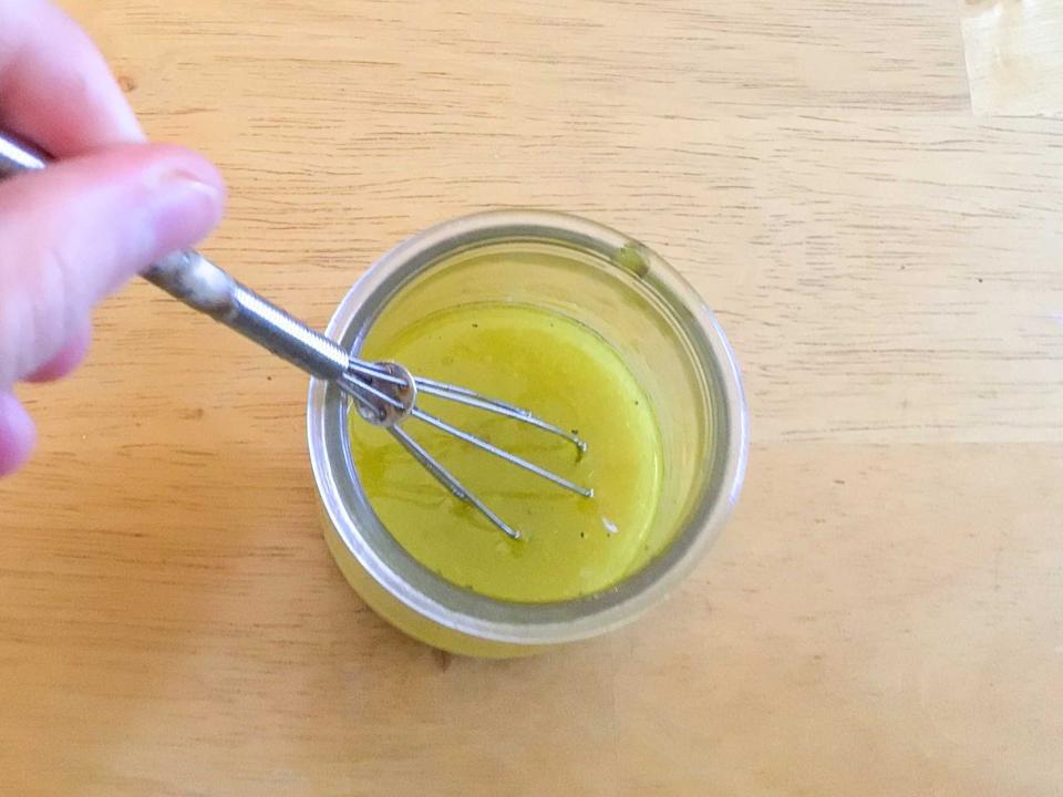 A jar of a yellow vinaigrette with a hand whisking the mixture inside the jar. The jar sits on a wooden cutting board