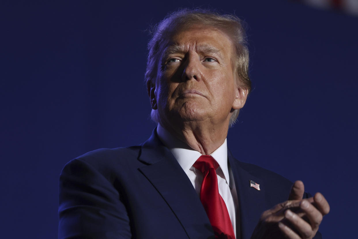 Former President Donald Trump, in his usual blue suit and red tie, claps.