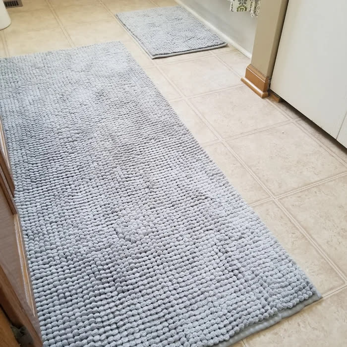 Reviewer's photo of two of the bath mats in different sizes, both light blue, on the floor in a bathroom, one in front of the sink and the other in front of the toilet