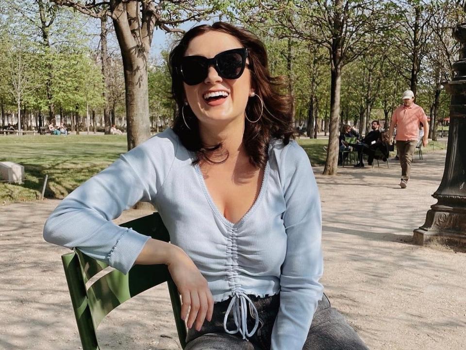 Ember Langley, a white woman in a blue top and sunglasses, laughs on a bench in a park in Paris.