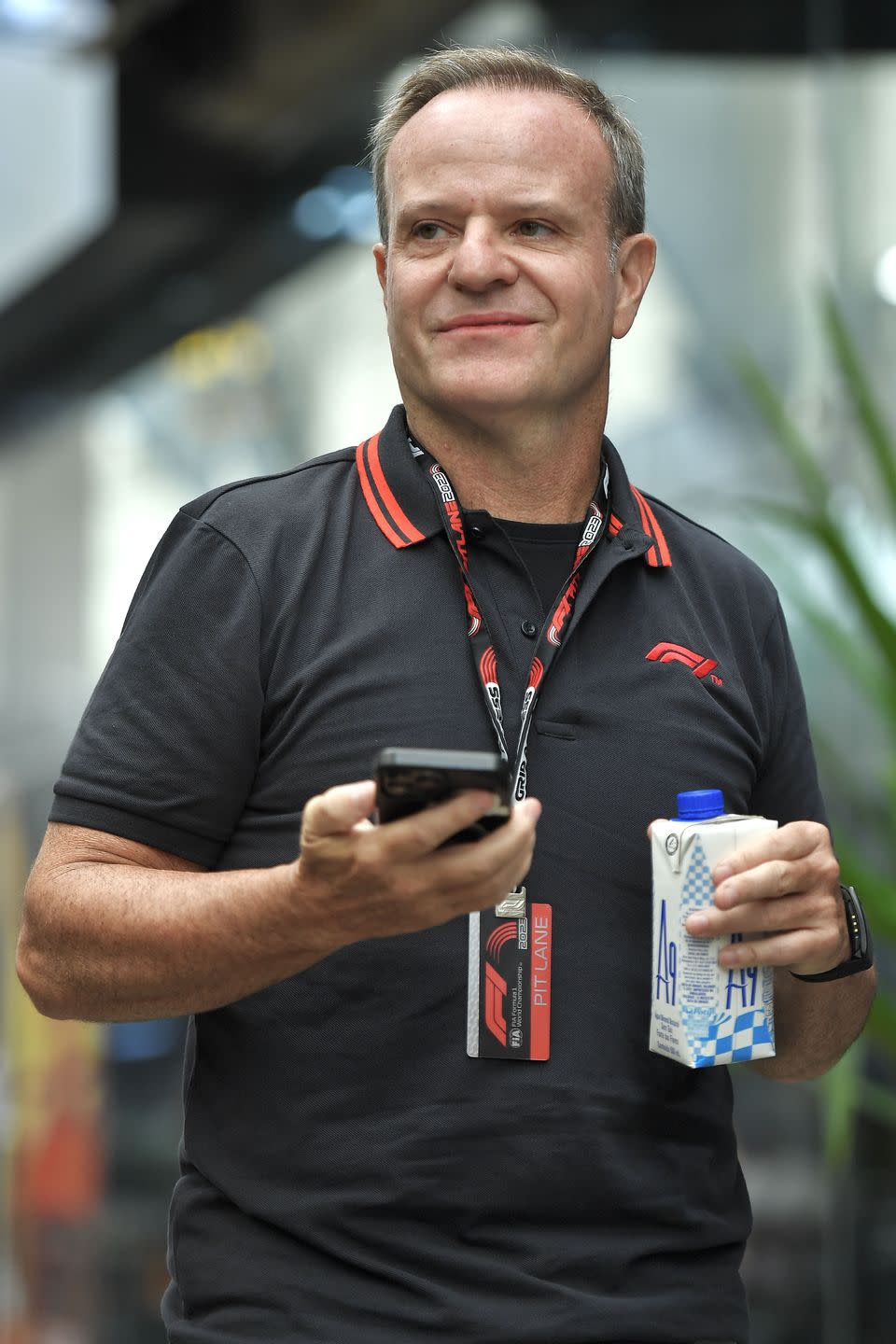 rubens barrichello at the 2023 brazilian grand prix, wearing f1 team kit and holding his phone in one hand and a drinks carton in the other hand