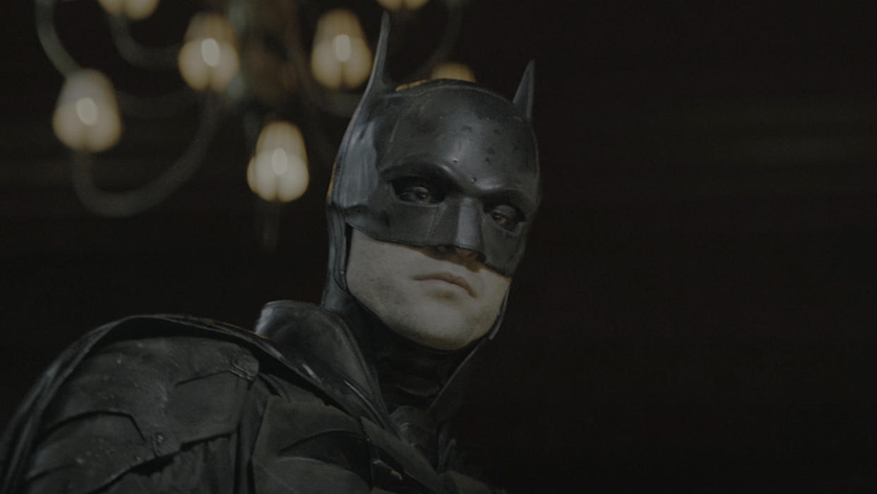  Robert Pattinson's Batman looking down at crime scene as camera flash goes off in The Batman. 