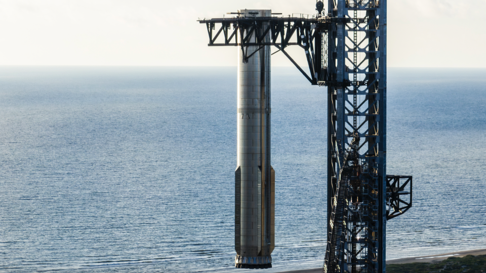     Nahaufnahme einer silbernen Trägerrakete, die von den mechanischen Armen ihres Startturms über dem Boden gehalten wird, mit dem Meer im Hintergrund. 