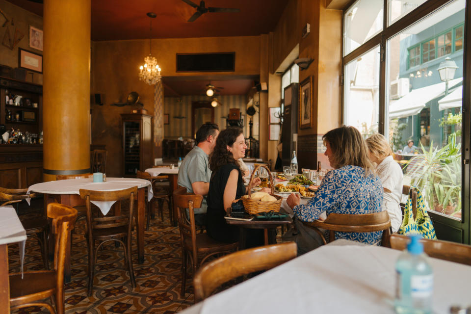 Four people dine at a cozy restaurant with large windows, surrounded by wooden furniture and décor with a vintage charm