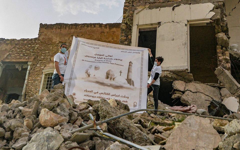 Youths unfurl a poster welcoming Pope Francis above the rubble of a destroyed house next to the ruins of the Syriac Catholic Church of the Immaculate Conception (al-Tahira) in the old city of Iraq's northern city of Mosul - AFP