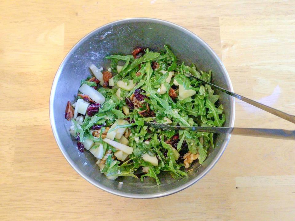 Bird's-eye-view of a metal bowl of a tossed salad containing arugula, cranberries, apple pieces, and nuts with metal tongs sticking out the bowl