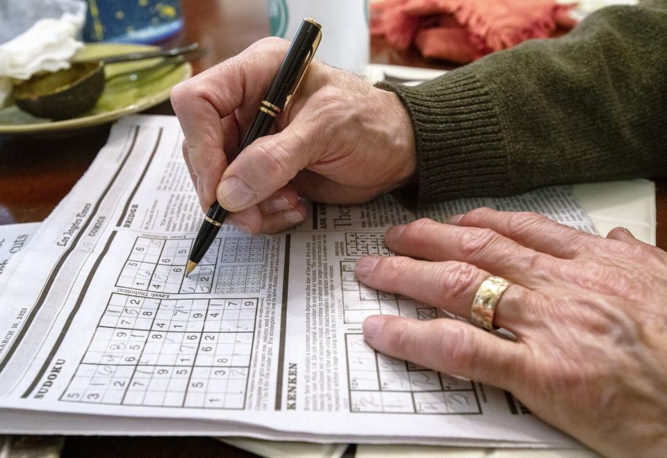 Hands rest on a newspaper page that displays crossword and Sudoku puzzles.