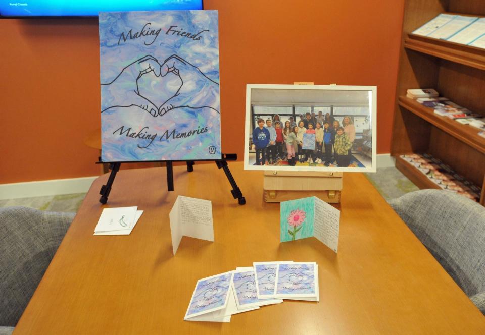 A display features the cover of a "Making Friends Making Memories" pen pal card created by Max Bohane, left, and a photo of the Plymouth River School fifth grade class members from Hingham, right, who are pen pals with residents at The Cordwainer in Norwell.