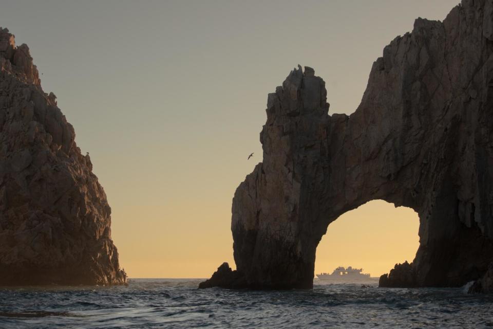 A boat on the water far away, framed at sunset by a large arched rock formation.