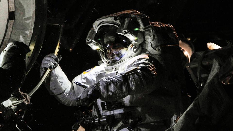     A haunting picture of astronaut Chris Cassidy in a space suit.  the astronaut holds the international space station patch with his right hand. 