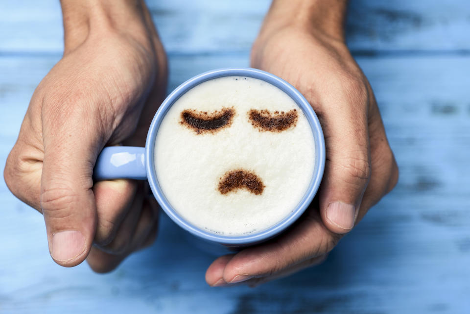 high-angle shot of a young caucasian man with a blue cup of cappuccino with a sad face drawn with cocoa powder on the milk foam, on a blue rustic table