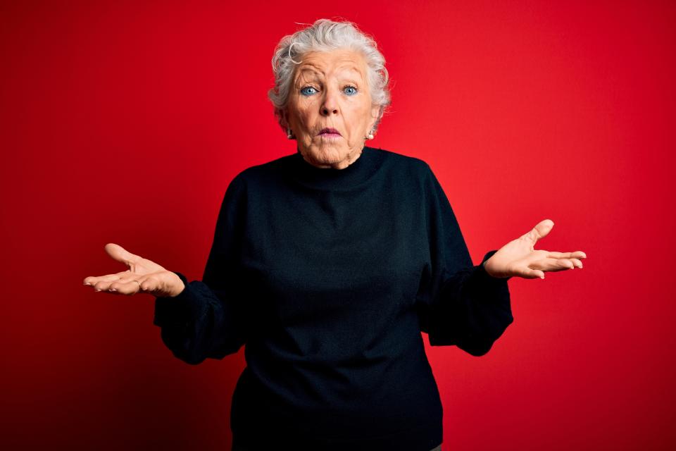 Older woman with short white hair wearing a black turtleneck, shrugging with her palms up against a plain background