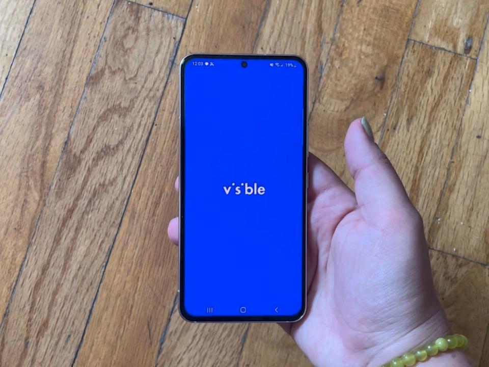 A hand holding a phone displaying the Visible home screen.