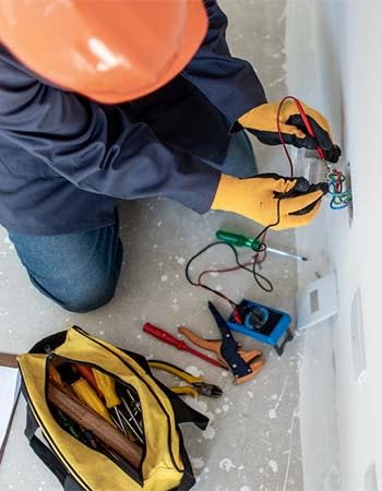 An electrician uses tools to assess electrical components in a wall.