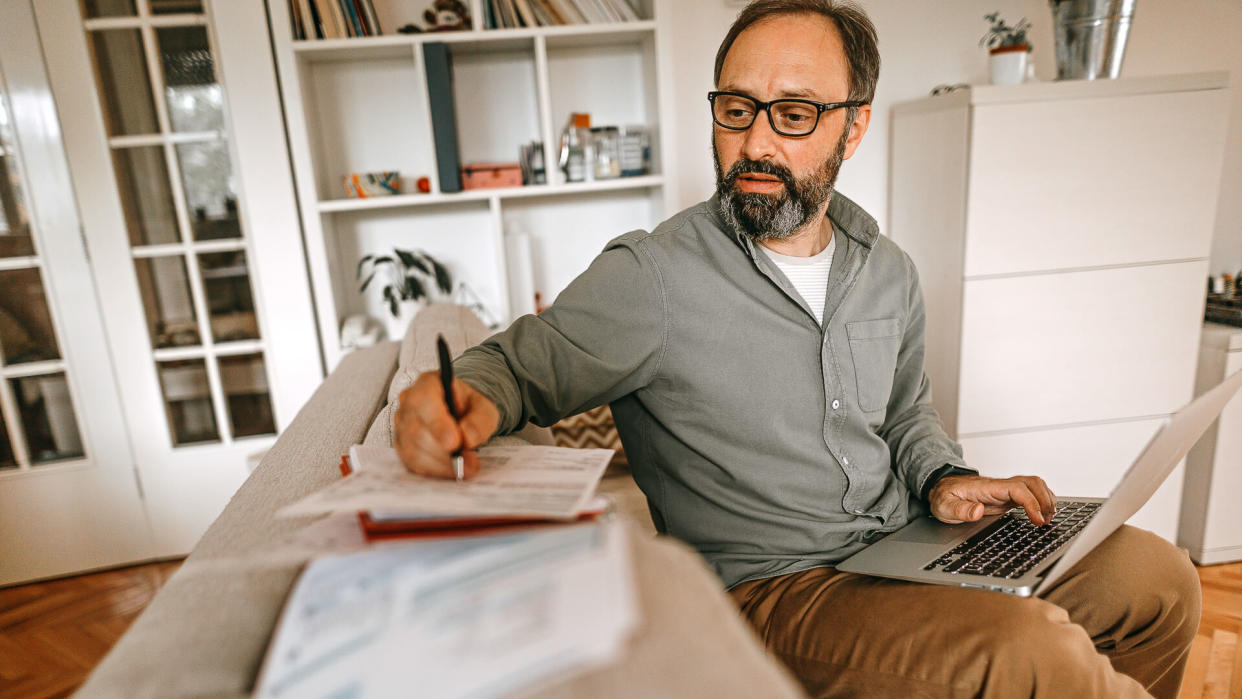 Mature man sitting on sofa in living room with laptop and financial reports, doing his monthly budget.