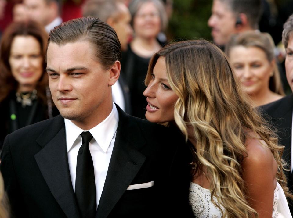 leonardo dicaprio, wearing a black suit and tie, and gisele bundchen, wearing a white dress, stand among a crowd and look off camera