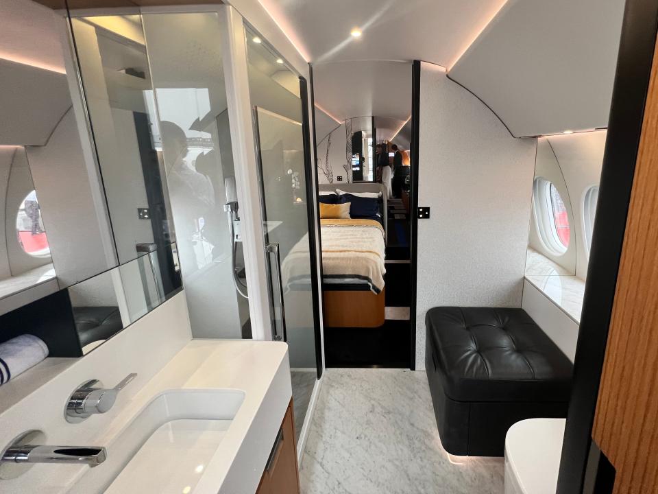 The bathroom of a Dassault Falcon 10X shows a sink, shower, toilet, and cushioned seat, with marble floor.