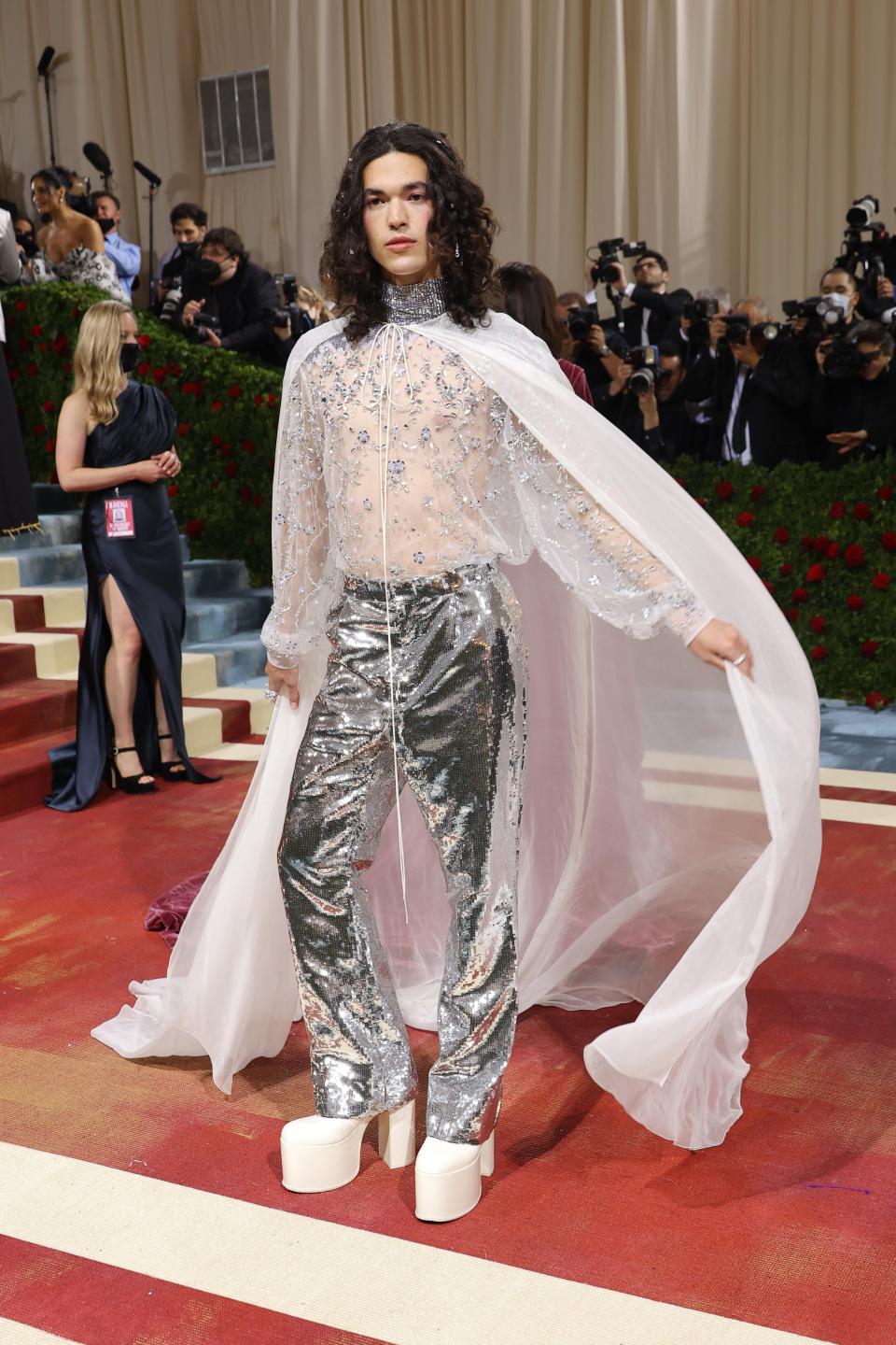 Conan in a sheer white blouse with silver embellishments throughout, silver metallic pants, tall white platform boots, and a sheer white billowing cape.