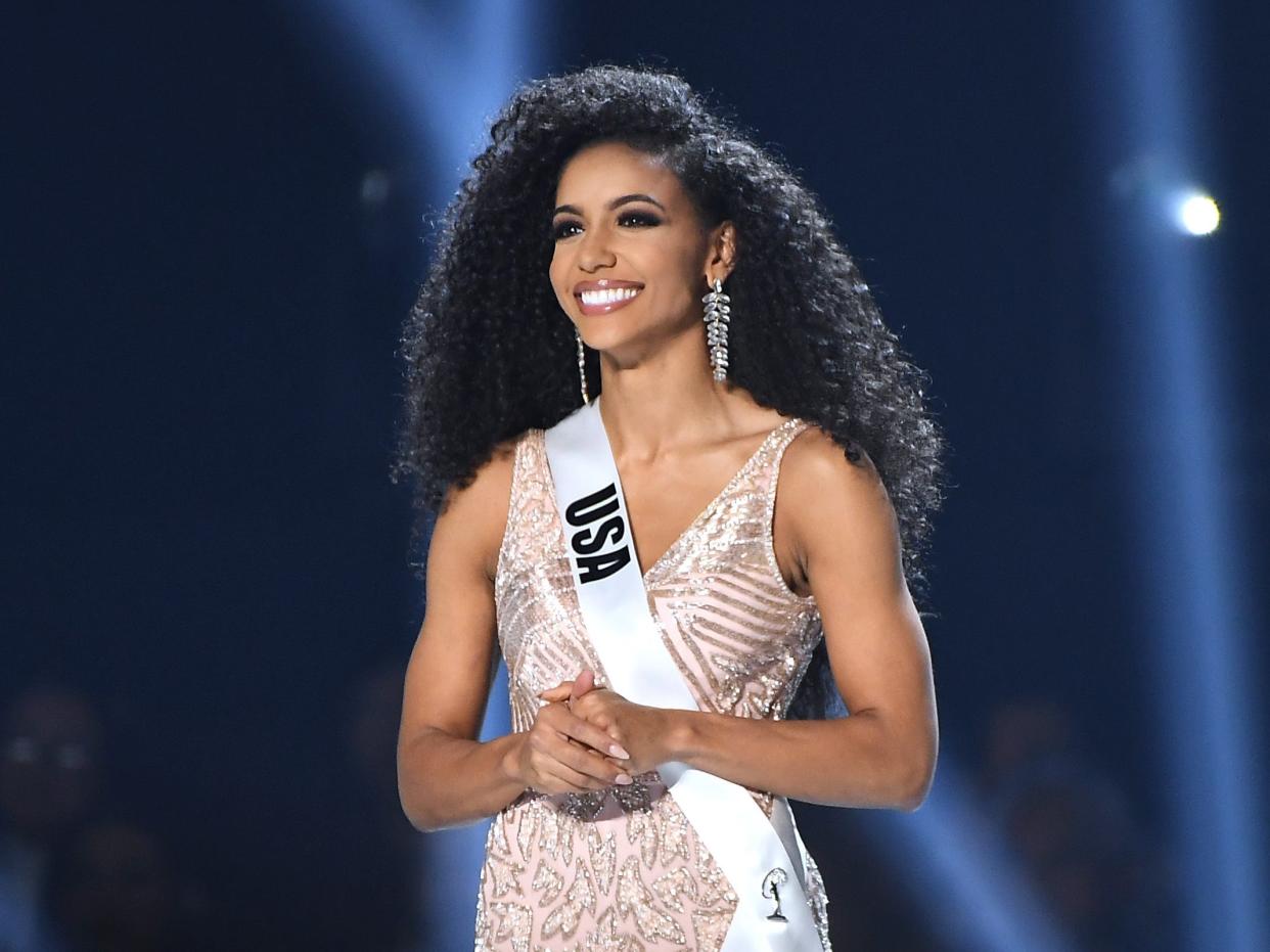 Cheslie Kryst onstage at the 2019 Miss Universe Pageant, spotlighted against a dark blue background.