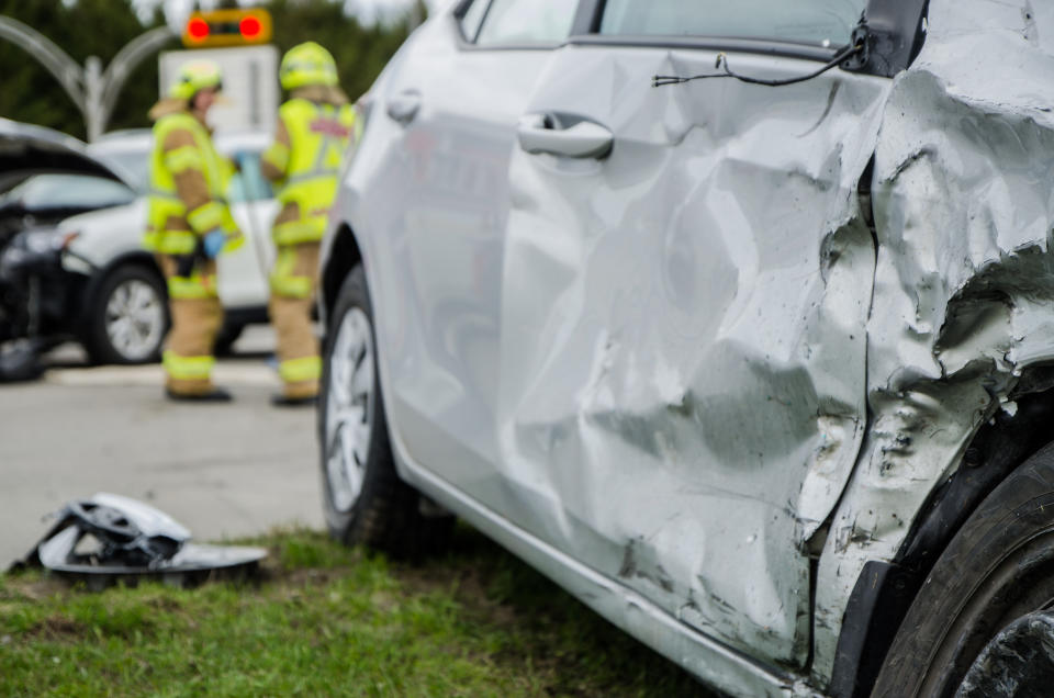 Two firefighters in protective gear are examining the scene of a car accident involving two heavily damaged vehicles