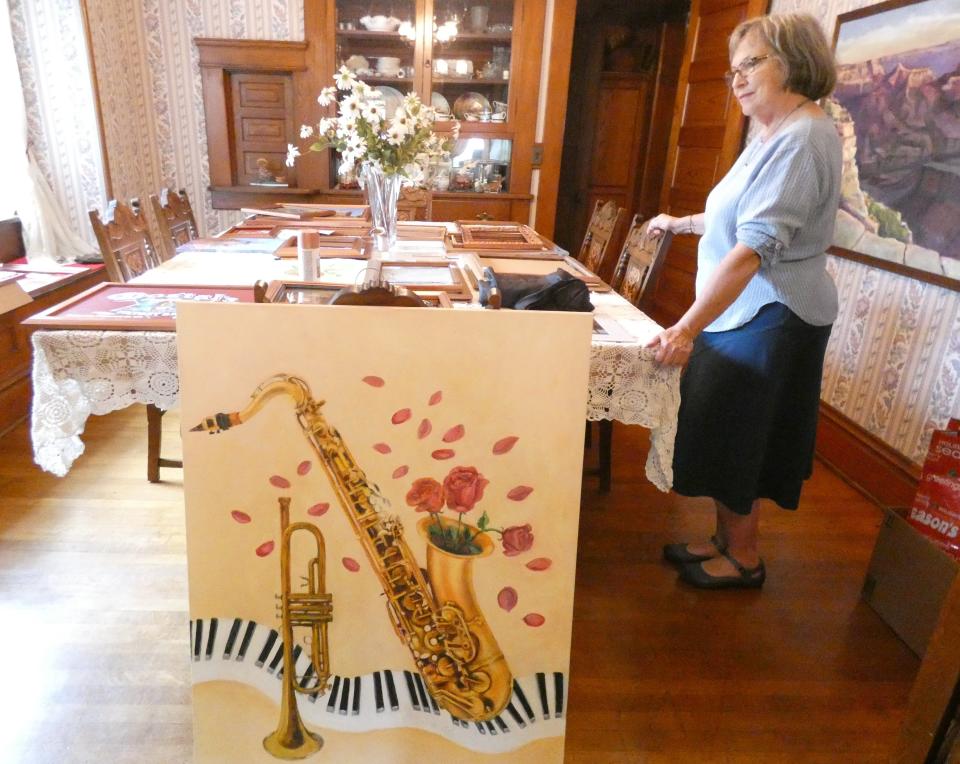 Artworks created for a special project showcasing Bucyrus history are arranged on Peg Vasil's dining room table. In the foreground is a painting by Vasil with a keyboard and some brass instruments that John Kennedy played, with roses coming out of the saxophone.