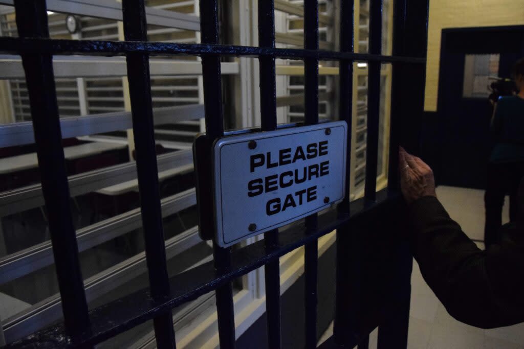 A hand holds a cell door with a white sign saying "Please secure gate."