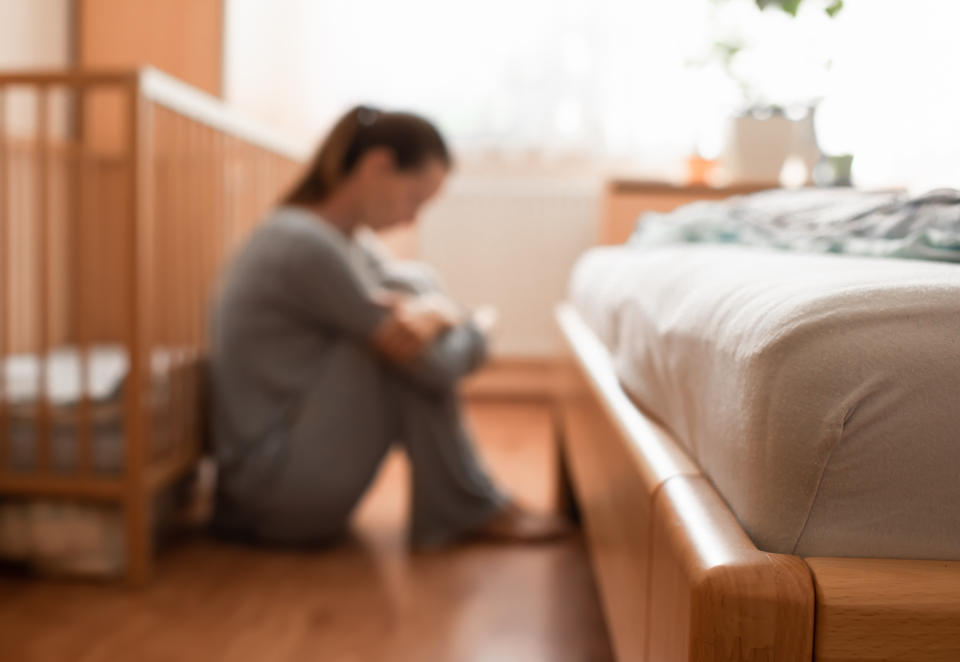 A woman sits on the floor next to a bed and a crib, hugging her knees and looking down, appearing pensive or stressed