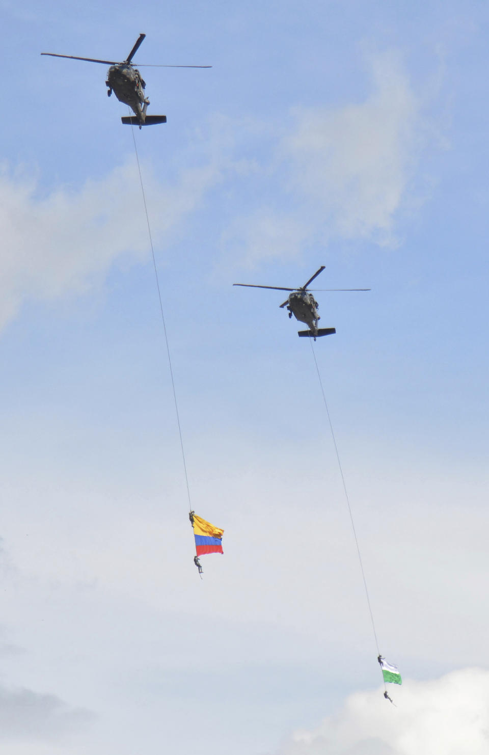 Two members of Colombia´s air force hang from a cable under a helicopter flying a Colombian flag, left, before plunging to their deaths when the cable snapped during the mid-air stunt at the Medellin Flower Fair in Medellin, Colombia, Sunday, Aug. 11, 2019. (AP Photo/Luis Benavides)