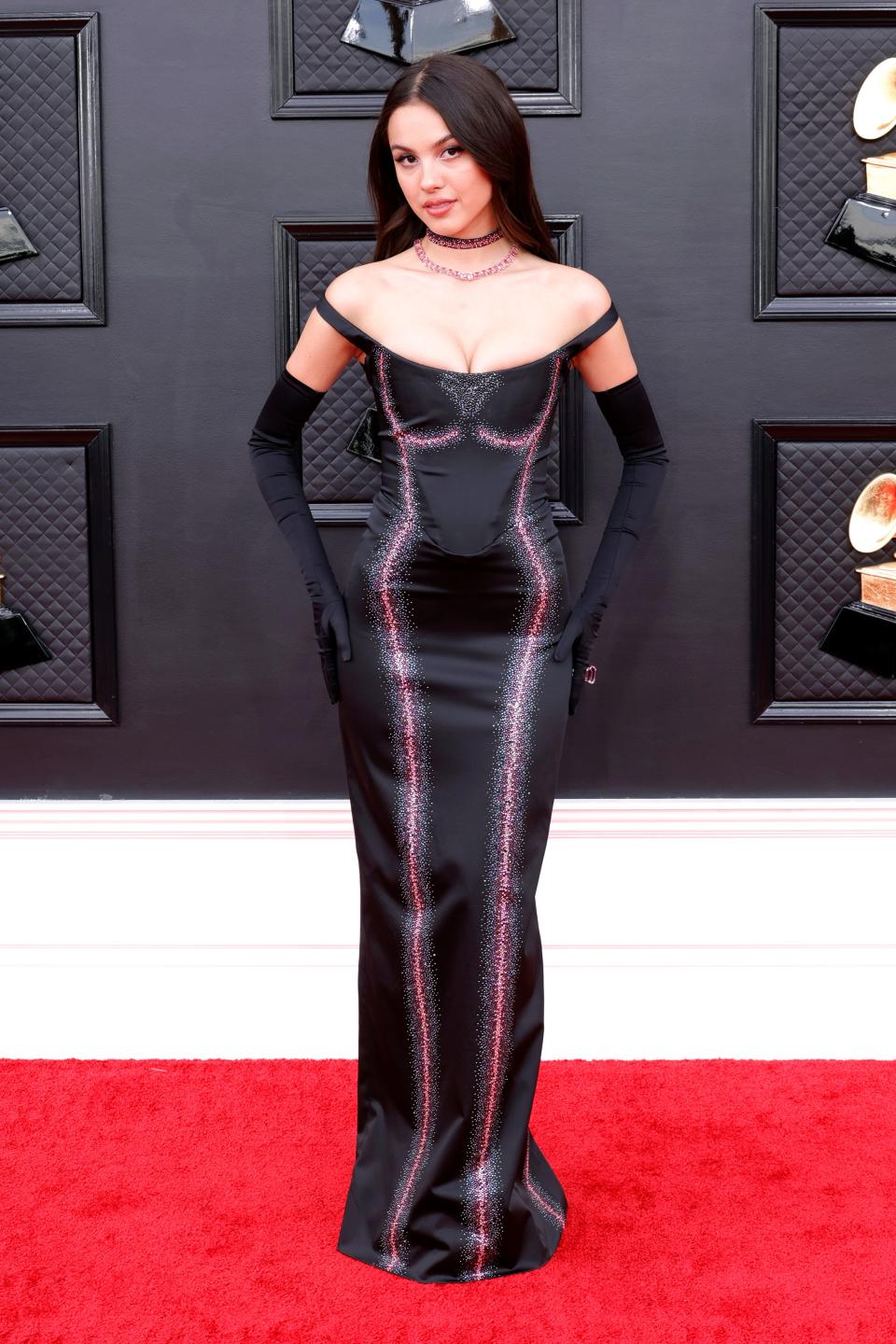 Olivia in a black off-the-shoulder satin gown showing off her cleavage. The dress has a faux outline of breasts and a silhouette of a body in pink sparkles. She's wearing black off the elbow gloves and two choker necklaces.