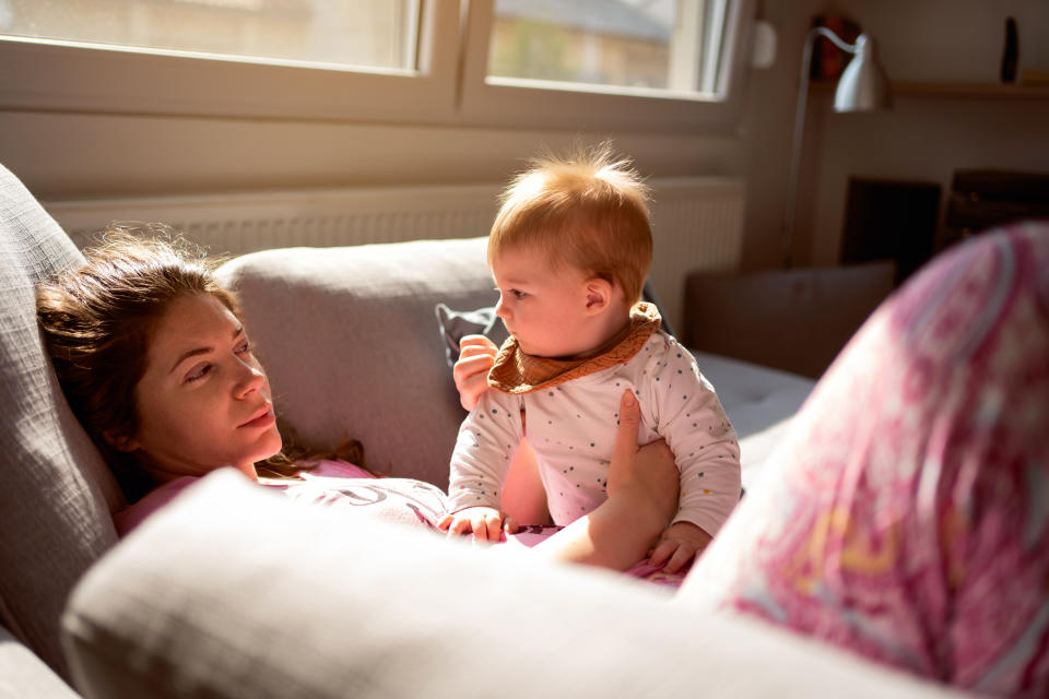 Caucasian tired mother laying down on the sofa and holding her baby son while they looking at each other face to face.