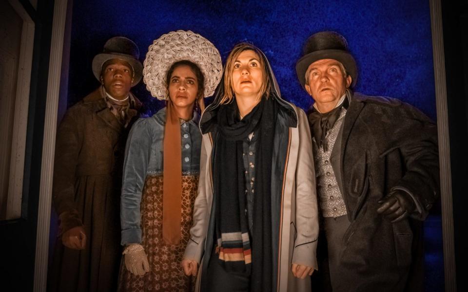 Ryan, Yaz, the Doctor, and Graham, all wearing period 1800s clothing, apart from the Doctor, stand soaked with rain in a brightly lit doorway in Doctor Who.