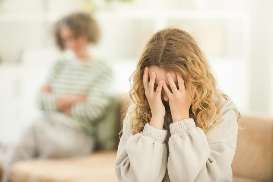 A young girl with her head in her hands as her mother sits cross-armed in the background