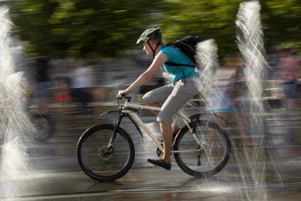 A man cools herself riding a bicycle through a fountain at a park in Moscow, Russia, Friday, Aug. 3, 2018. The hot weather in Moscow is continuing, with temperatures forecast to reach 30 degrees Celsius (86 Fahrenheit) . (AP Photo/Alexander Zemlianichenko)