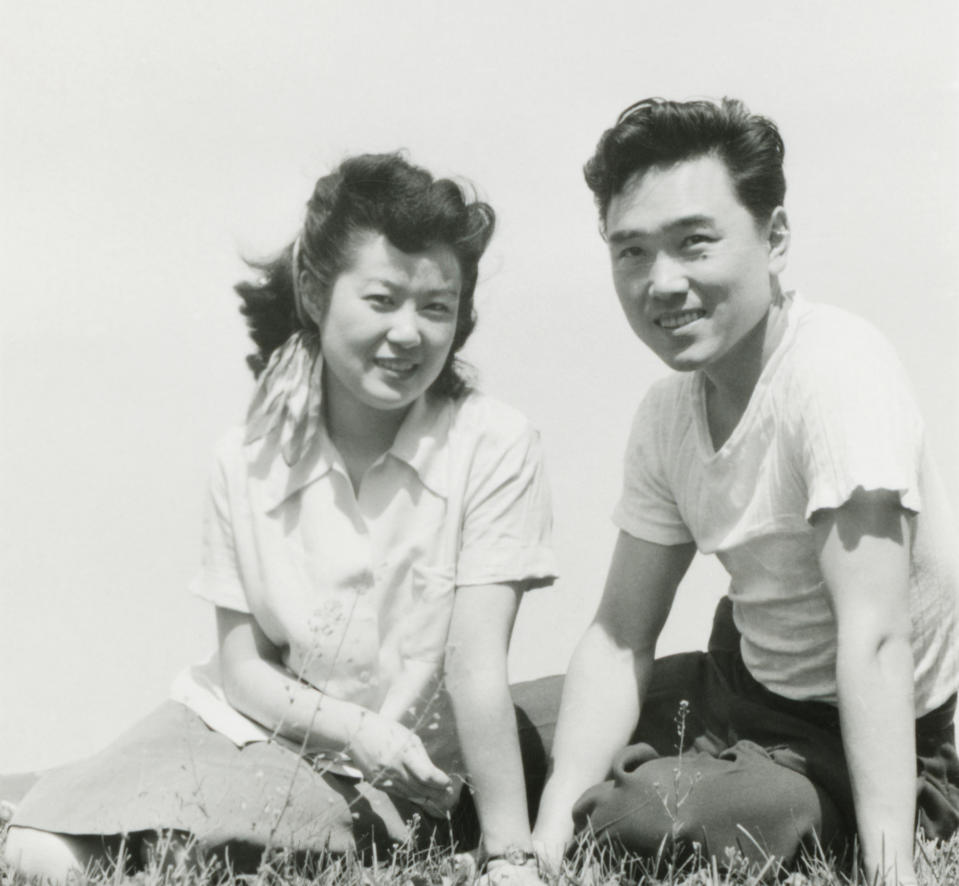 A man and a woman sit closely together on the grass, smiling; both wearing casual clothing. Names not provided