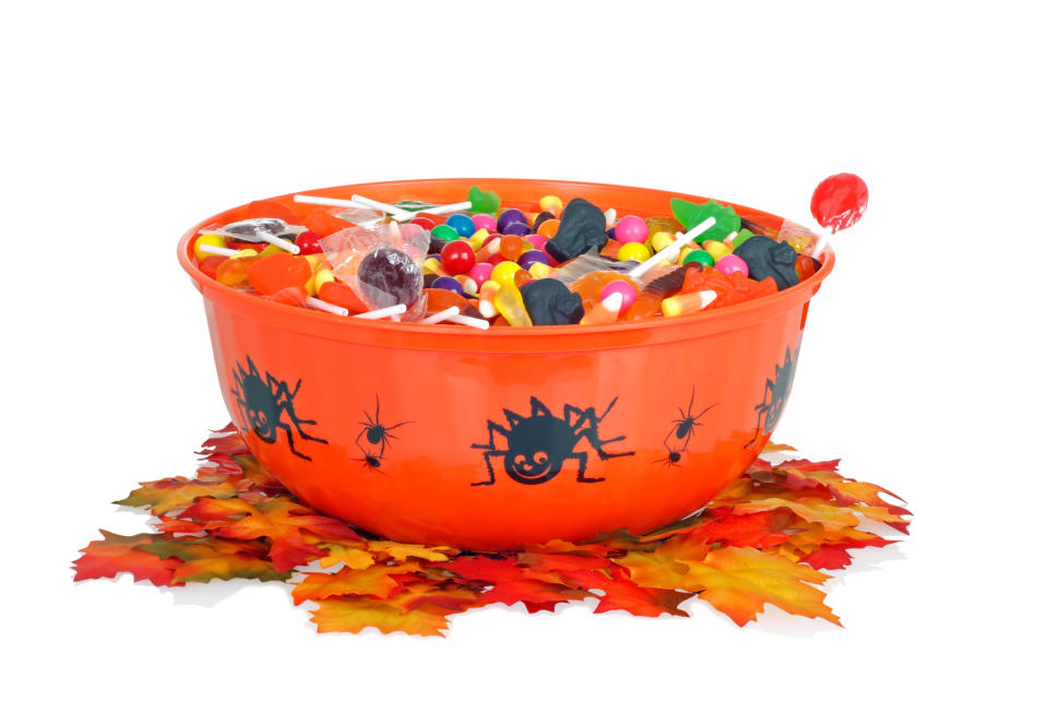 A orange bowl with spider design is filled with assorted Halloween candies, placed on a bed of autumn leaves