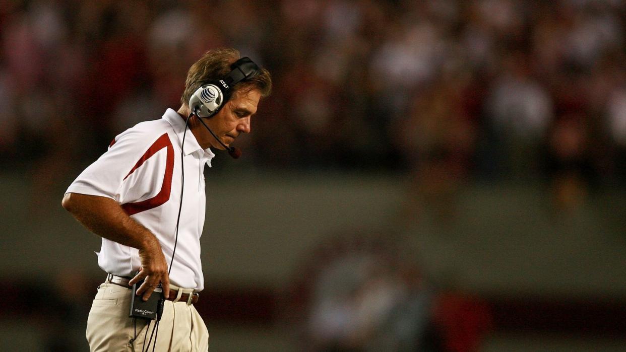 nick saban looks down and walks to the right, he wears a white polo shirt and khakis with a headset, he adjusts the headset receiver on his belt with on hand