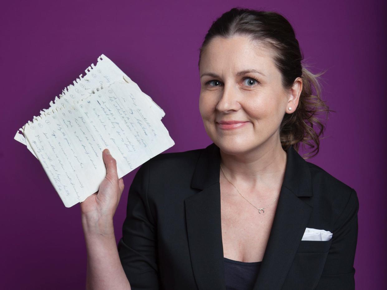 Laura Carney sits, smiling slightly at the viewer and holding several hand-written pages in her right hand, while her left rests in her lap. She wears a black blazer with a white handkerchief in the pocket, a black tank top, and black pants. She has blues eyes and dark blonde hair pulled back into a ponytail. The background is an eggplant purple.