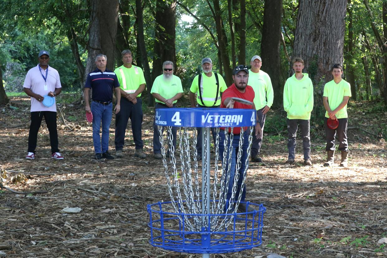 With the crew that helped build the course behind him, City of Zanesville Supervisor for Sewer and and Storm Sewer Maintenance Chris Fleming throws a putt at the new disc golf course at Riverside Park.